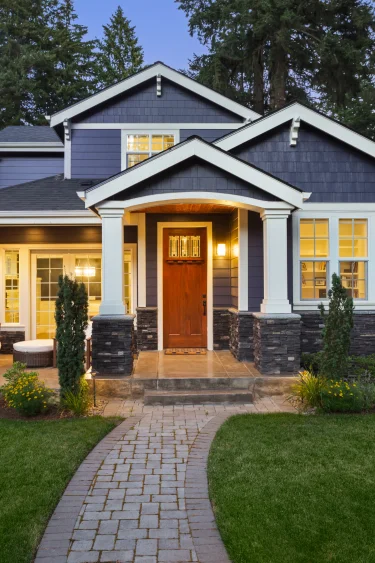 A dark blue house with white trim stands against a backdrop of trees. The house features a wooden front door, accessible via a paved walkway that cuts through a green lawn.