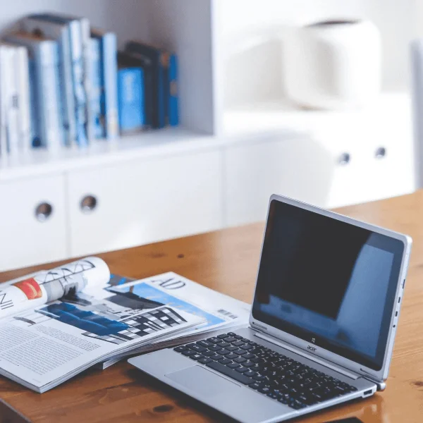 An open laptop sitting on a wooden table next to an open magazine in a well-lit home office.