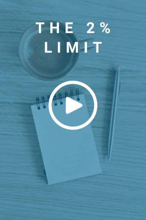 Aerial view of a cup of coffee, a pen, and an open notepad all placed on a wooden desk. There is a text overlay that reads 'The 2% Limit'.